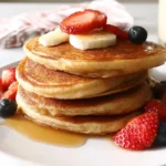 Fluffy pancakes from a keto pancake recipe with coconut flour, stacked and topped with banana slices, strawberries, and blueberries, served with syrup on a white plate.