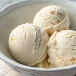 ultra-close shot of keto cottage cheese ice cream. Overhead close‑up of three ice‑cream scoops in a wide, shallow white ceramic bowl; the central scoop is slightly higher, all scoops have a smooth glossy surface with swirl marks